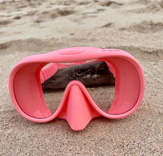 A pink dive mask with a clear vision lens and silicone nose piece lying on sandy beach.