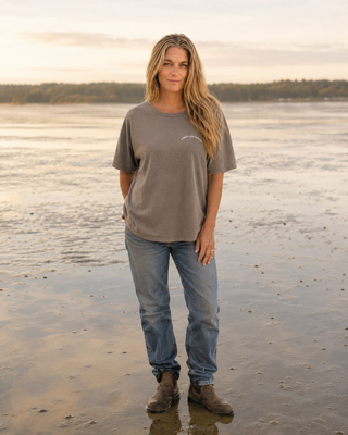 A woman stands on a wet shoreline at sunset wearing an oversized taupe-gray t-shirt with a small white script logo on the left chest, blue jeans, and brown boots.