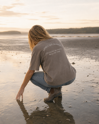 A woman crouches on a wet beach at sunset wearing a taupe gray t-shirt with 'APRÈS SEA RANCH' printed on the back, paired with blue jeans and brown boots.
