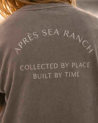 Close-up of the back of a person wearing a faded dark gray t-shirt with white text reading 'APRÈS SEA RANCH' and 'COLLECTED BY PLACE BUILT BY TIME'.