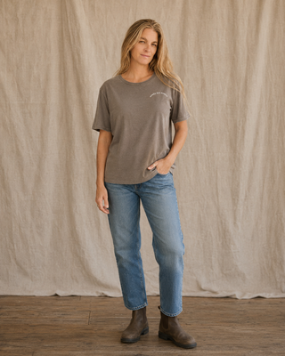 A woman stands in front of a beige fabric backdrop wearing a muted taupe short-sleeve t-shirt with a small white graphic on the left chest, blue jeans, and dark brown boots.