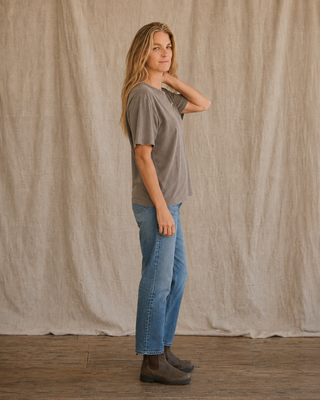 A woman stands in profile wearing a loose taupe-gray short-sleeve t-shirt, blue jeans, and brown ankle boots, posed against a draped beige fabric backdrop.