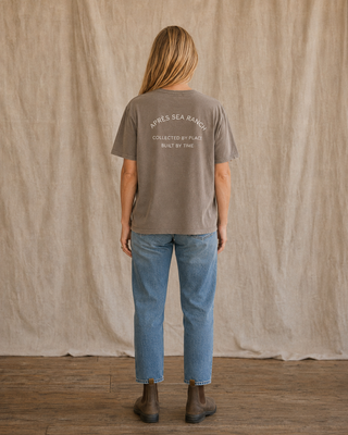 A woman stands with her back to the camera wearing a faded taupe crewneck t-shirt with white text on the back reading 'APRÈS SEA RANGE COLLECTED BY PLACID BUILT BY TIME', paired with blue jeans and brown boots, set against a neutral beige fabric backdrop.