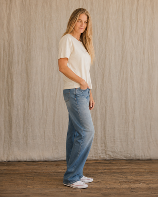 A woman stands in profile wearing a cream short-sleeve crewneck t-shirt, light-wash wide-leg blue jeans, and white slip-on sneakers, posed against a textured beige fabric backdrop on a wooden floor.