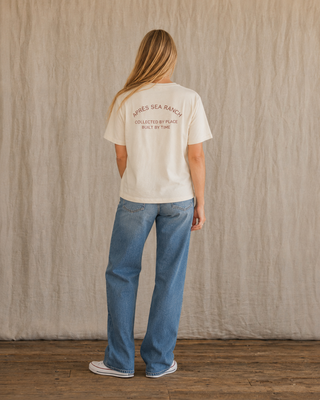 A woman stands with her back to the camera wearing a cream-colored t-shirt with 'SIRIES SEA RANCH' printed in brown on the back, paired with blue jeans and white sneakers, set against a neutral beige fabric backdrop.