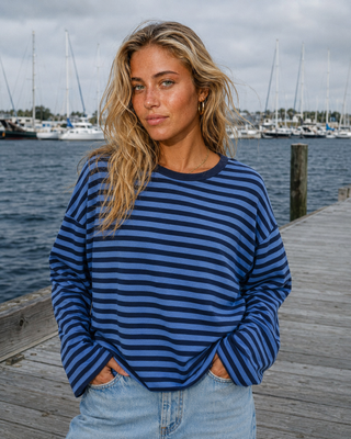 A woman with blonde hair stands on a wooden dock wearing a blue and navy striped long-sleeve top and light wash jeans, with a marina and sailboats in the background.