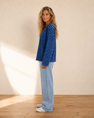 A woman stands in profile wearing a blue and black striped long-sleeve knit top, light-wash wide-leg jeans, and white sneakers against a warm beige wall and light wood floor.