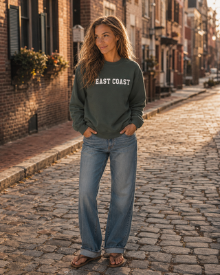 A woman stands on a cobblestone street wearing a dark olive green sweatshirt with 'EAST COAST' printed in white across the chest, paired with light blue jeans and brown sandals.