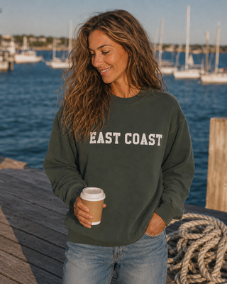 A woman standing on a wooden dock by the water wearing an olive green sweatshirt with 'EAST COAST' printed in white across the chest, holding a coffee cup.