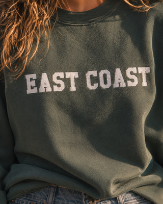 A close-up of a person wearing a dark olive green crewneck sweatshirt with 'EAST COAST' printed across the chest in white varsity-style letters, paired with blue jeans.