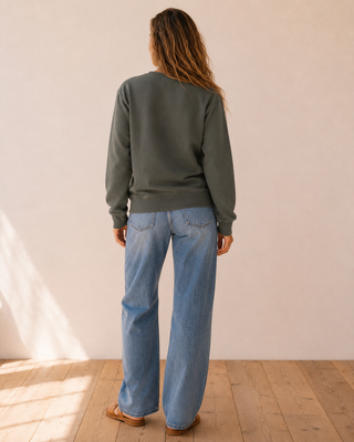 A woman viewed from behind wearing a dark olive green crewneck sweatshirt and light blue wide-leg jeans, standing on a light wood floor against a neutral beige wall.