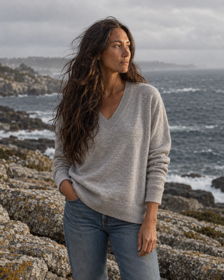 A woman with long dark hair stands on rocky coastal ground wearing an oversized light gray V-neck sweater and blue jeans, with the ocean and cloudy sky in the background.