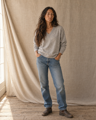 A woman stands indoors wearing a light gray V-neck sweater, blue jeans, and brown boots, with her hands in her pockets, posed in front of a draped beige fabric backdrop.