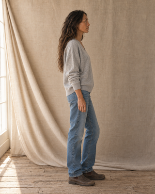 A woman stands in profile wearing a light gray long-sleeve knit sweater, blue straight-leg jeans, and brown boots, posed in front of a draped beige fabric backdrop.
