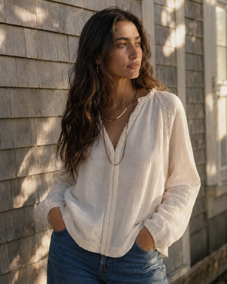 A woman wearing a cream-colored, textured long-sleeve blouse with a split V-neckline, paired with blue jeans, standing against a gray wooden shingled wall.