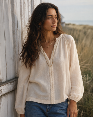 A woman wearing a cream-colored, long-sleeve linen blouse with lace trim and a V-neckline, paired with blue jeans, standing outdoors next to a weathered white wooden fence.