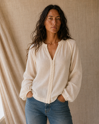 A woman wearing a cream-colored, long-sleeve textured blouse with decorative trim down the front and gathered cuffs, paired with blue jeans, standing against a neutral beige linen backdrop.