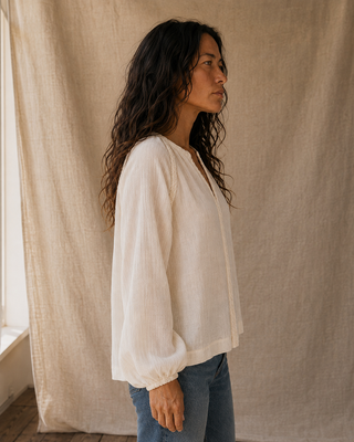 A woman with long, wavy dark hair is shown in profile wearing a loose, long-sleeve white blouse and blue jeans, standing in front of a draped beige linen backdrop.