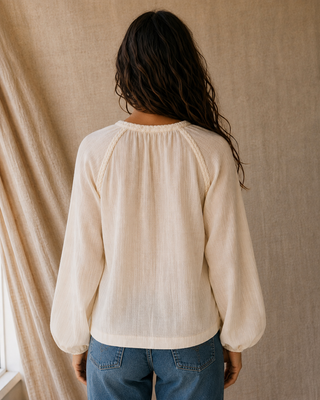 A woman viewed from behind wearing a cream-colored long-sleeve blouse with a gathered neckline and balloon sleeves, paired with blue jeans, standing against a neutral linen-textured backdrop.