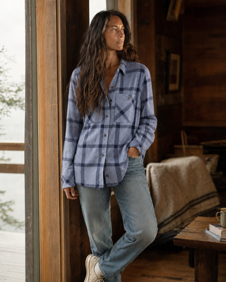 A woman stands in a rustic cabin interior wearing a blue and gray plaid button-up flannel shirt, light-wash jeans, and white sneakers.