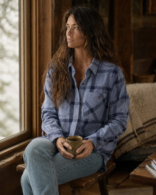 A woman with long dark hair sits on a wooden stool by a window, wearing a blue plaid button-up shirt and light blue jeans, holding a small ceramic mug.