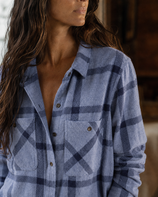 A close-up of a woman wearing a light blue and navy plaid button-up shirt. The photo focuses on the texture of the fabric and the plaid pattern, with the model's face partially cropped on the left.