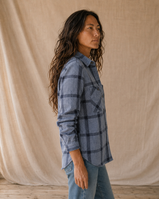 A woman with long dark hair is shown in profile wearing a blue-gray and navy plaid button-up shirt with blue jeans, standing in front of a draped beige fabric backdrop.