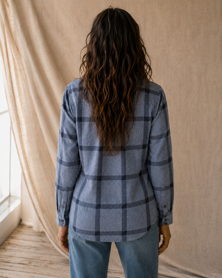 A woman viewed from behind wearing a slate blue and black plaid flannel shirt, paired with light blue jeans, standing in front of a draped beige fabric backdrop.