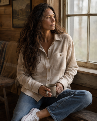 A woman with long dark hair sits on a wooden bench in a rustic room, wearing a light beige long-sleeve button-up shirt, blue jeans, and white sneakers while holding a ceramic mug.