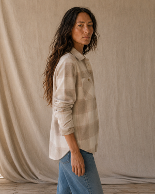 A woman wearing a beige and off-white plaid flannel shirt with blue jeans, standing in front of a draped beige fabric backdrop.