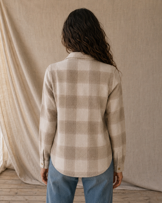 A woman viewed from behind wearing a beige and cream plaid flannel shirt and blue jeans, standing in front of a draped linen backdrop.