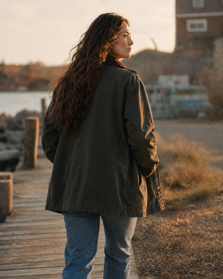 A woman with long, wavy brown hair stands on a wooden boardwalk at sunset, viewed from behind as she wears an oversized dark olive green jacket and blue jeans.