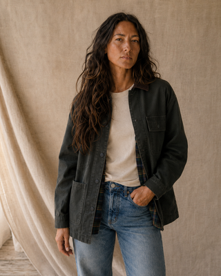A woman wearing a dark charcoal utility overshirt over a cream t-shirt and blue jeans, standing against a draped beige linen backdrop.