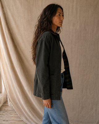 A woman wearing a dark charcoal utility jacket over a black-and-white plaid button-up shirt and light blue jeans, standing in profile against a draped beige linen backdrop.