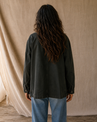 A person viewed from behind wearing a dark charcoal gray oversized jacket and light blue jeans, standing in front of a draped beige fabric backdrop.