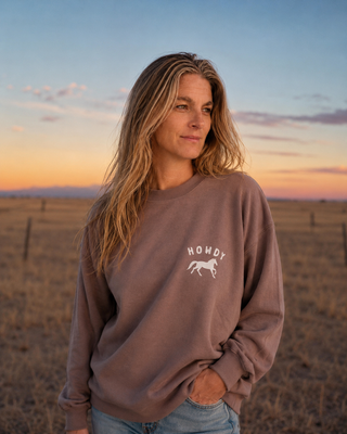 A woman standing in a dry field at sunset wearing a taupe crewneck sweatshirt with a white graphic that reads 'HOWDY' and a horse silhouette, paired with light blue jeans.