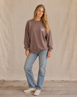A woman modeling a brown crewneck sweatshirt with a white graphic print on the left chest, paired with light blue jeans and white sneakers, standing against a neutral beige fabric backdrop.