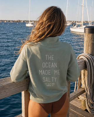 A woman seen from behind wearing a sage green quarter-zip sweatshirt with white text on the back that reads 'THE OCEAN MADE ME SALTY' and 'THE SALT LIFE KEMPER, OH'. She is standing on a wooden dock with the ocean and sailboats in the background.