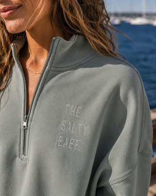 A close-up of a woman wearing a sage green half-zip sweatshirt with 'THE SALTY BABE' embroidered on the chest, set against a sunny outdoor background with blue water and boats.