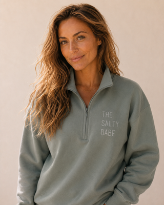 A woman with long wavy brown hair wearing a sage green quarter-zip sweatshirt with the text 'THE SALTY BABE' printed in white on the left chest, set against a plain beige background.