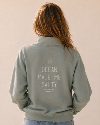A woman viewed from behind wearing a sage green sweatshirt with the text 'THE OCEAN MADE ME SALTY' printed in white on the back, paired with light blue jeans against a solid beige background.