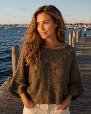 A woman wearing a chunky brown knit sweater with wide three-quarter sleeves, standing on a wooden dock with water and boats in the background.