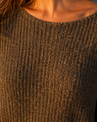 Close-up of a person wearing a ribbed knit sweater in a warm brown or taupe color, with the neck and upper chest visible against warm golden light.