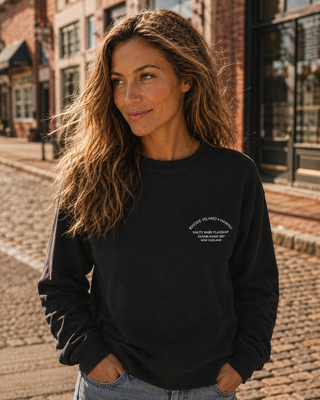 A woman with long wavy brown hair stands on a cobblestone street wearing a black crewneck sweatshirt with white text on the left chest that reads 'Salty Babe Flagship. Rhode Island Hawaii .