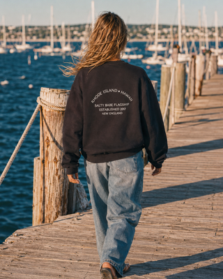 A person viewed from behind walking on a wooden dock by the water, wearing a black sweatshirt with white printed text on the back reading 'RHODE ISLAND + HAWAII SALTY BABE FLAGSHIP ESTABLISHED 2017 NEW ENGLAND' and light blue jeans.