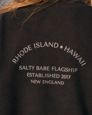 Close-up of a black sweatshirt featuring white printed text on the back that reads 'RHODE ISLAND • HAWAII', 'SALTY BABE FLAGSHIP', 'ESTABLISHED 2017', and 'NEW ENGLAND'.