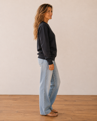A woman in side profile wearing a dark charcoal crewneck sweatshirt and light-wash wide-leg jeans, standing on a wooden floor against a plain beige wall.