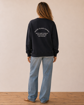 A woman stands with her back to the camera wearing a black crewneck sweatshirt with white text on the back reading 'RHODE ISLAND + HAWAII' and 'SALTY BABE FLAGSHIP ESTABLISHED 2017 NEW ENGLAND', paired with light blue jeans.