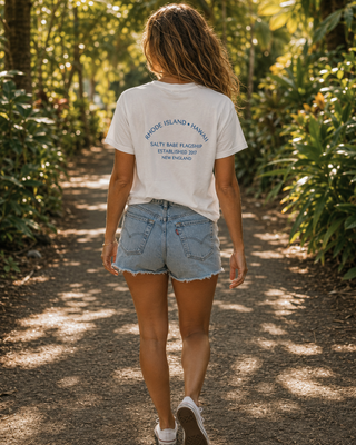 A woman walking away on a sun-dappled forest path, wearing a white graphic T-shirt with blue text reading 'BAYONDE ISLAND YOUTH' and 'SILKY RABBIT FOUNDATION EST. 2021', paired with light-wash denim shorts and white sneakers.