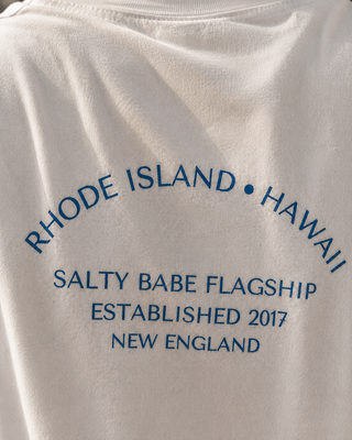Close-up of a white T-shirt with blue printed text reading 'RHODE ISLAND • HAWAII', 'SALTY BABE FLAGSHIP', 'ESTABLISHED 2017', and 'NEW ENGLAND'.